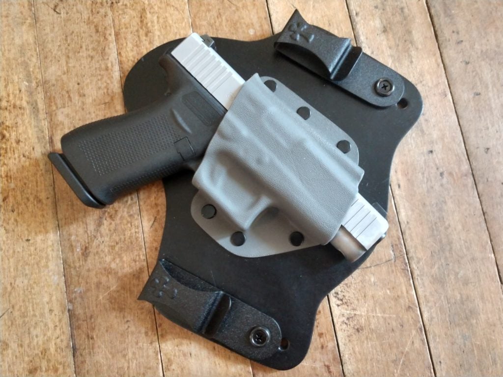 A two-tone black and silver Glock G48 holstered inside a gray Kydex and black leather hybrid CrossBreed SuperTuck inside-the-waistband (IWB) holster that is lying on a rustic hardwood floor.