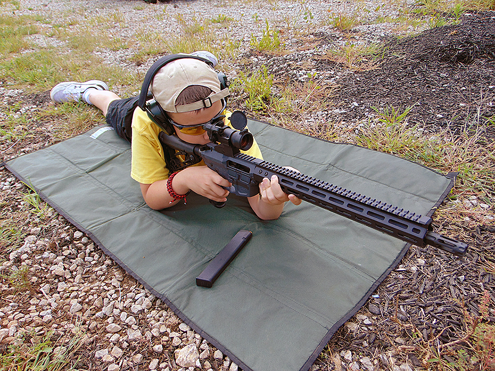 The author's son aims down the sights of a Wilson Combat AR-9 carbine while lying on a sniper mat at an outdoor range.