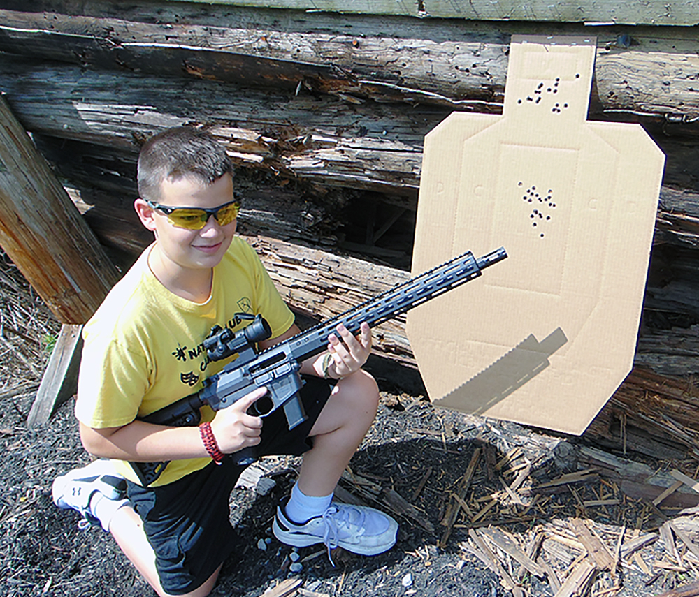 The author's son smiles as he kneels beside a cardboard target which contains several bullet holes. The young man is cradling a Wilson Combat AR-9 carbine.